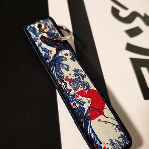 May include: Embroidered rectangular keyring featuring a Japanese wave and mountain design with a red sun, blue waves, and floral accents. The keyring has a navy blue border and a silver key ring.