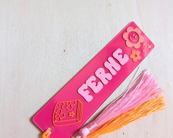 May include: A rectangular hot pink acrylic bookmark featuring the name FERNE in light pink bubble letters. The design includes an orange flower with a smiley face, small stars, and a book icon with floral details. A pink and orange tassel is attached.
