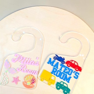 May include: Two clear acrylic door hangers. The left hanger features the pink text "Mila's Room" with a mermaid tail, starfish, and seashell. The right hanger features the blue text "MATEO'S ROOM" with a blue motorcycle, red car, yellow truck, and green car.