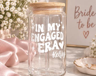 Könnte beinhalten: Ein durchsichtiges Glas mit Bambusdeckel und Glasstrohhalm. Auf der Vorderseite befindet sich ein weißer Schriftzug "IN MY ENGAGED ERA" mit kleinen Stern- und Herzsymbolen, gefolgt von dem Namen "Kelsey" in einer Schreibschrift unter dem Text.