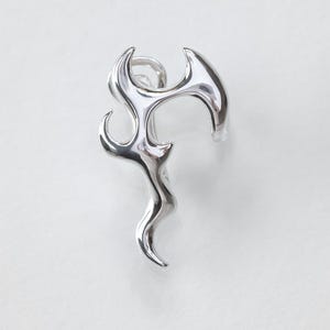Può includere: Un ear cuff color argento caratterizzato da una forma organica e fluida che ricorda una fiamma stilizzata o una forma liquida astratta. Il pezzo presenta una superficie metallica lucida e riflettente con bordi lisci e arrotondati.