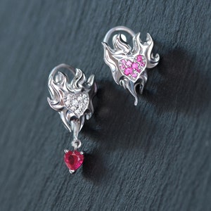 May include: A pair of silver-toned earrings featuring a flame-shaped design. One earring has a heart-shaped center with clear stones, while the other has a heart-shaped center with pink stones. A small red heart-shaped charm hangs from the flame base.