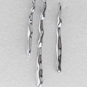 May include: A pair of silver-toned earrings featuring a fluid, molten metal design. The larger earring has an inverted U-shape with two long, dangling, wavy extensions, while the smaller earring is a single, elongated, irregular vertical drop.