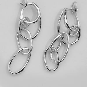 May include: A pair of silver-toned drop earrings featuring a series of interconnected, irregular, organic-shaped links. Each earring consists of three distinct, textured loops that hang vertically from a secure hinged hoop closure mechanism.