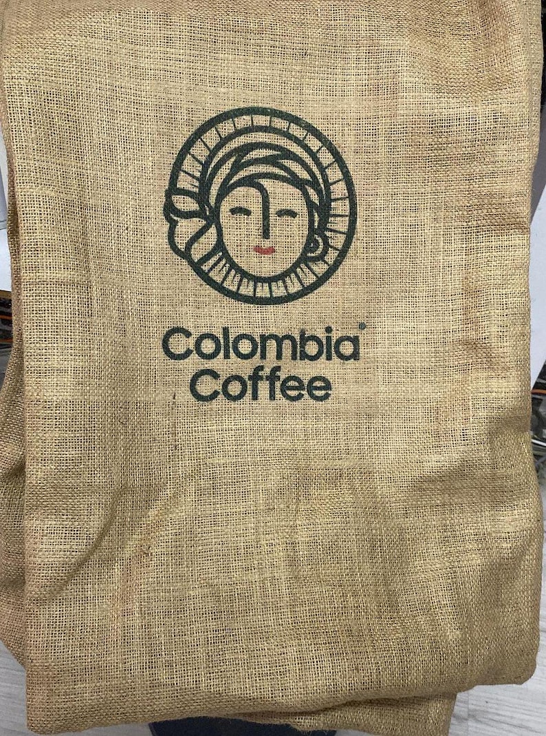 May include: A brown burlap sack with a black and white logo of a woman's face with the text "Colombia Coffee" printed on it.