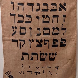 May include: A burlap fabric wall hanging with the Hebrew alphabet printed in black. The letters are in a traditional style and are arranged in rows. The bottom of the hanging features a black handprint and braille letters.