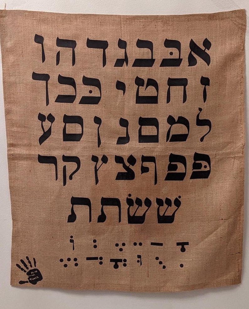 May include: A burlap fabric wall hanging with the Hebrew alphabet printed in black. The letters are in a traditional style and are arranged in rows. The bottom of the hanging features a black handprint and braille letters.