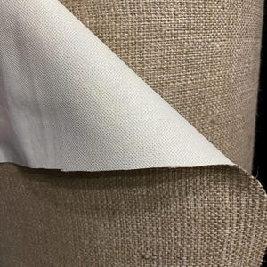 May include: Close-up of a roll of brown burlap fabric with a white backing. The burlap has a woven texture and the backing is smooth.