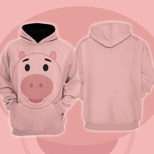 Pode incluir: Um moletom rosa claro com um rosto de porco de desenho animado na frente, com olhos pretos e um focinho rosa. O capuz tem um forro preto. A parte de tr&aacute;s do moletom &eacute; rosa claro liso. O moletom tem um bolso canguru.