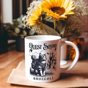 May include: White ceramic mug with a black and white illustration of a cat with a backpack, a castle, and the words "Quest Squad" and "Broccoli". The mug has a large handle and is set on a light brown surface.