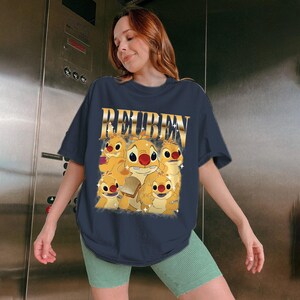 May include: Navy blue t-shirt featuring the word "REUBEN" in gold lettering above a graphic of several cartoon characters. The characters are yellow with red noses and are surrounded by stars. The shirt is worn by a person.