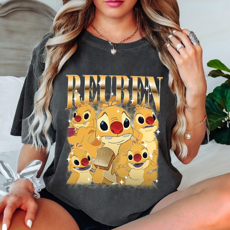 May include: Dark gray t-shirt featuring a graphic of the character Reuben from the animated film Lilo & Stitch. The design includes the word "Reuben" in gold lettering above a group of cartoon characters in shades of yellow and orange.