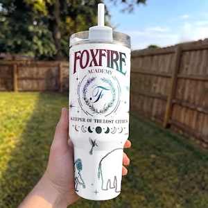 May include: White tumbler with the text "FOXFIRE ACADEMY" in a gradient of purple and teal. The tumbler features a design with a "T" inside a wreath, crescent moons, stars, and a hummingbird. The words "KEEPER OF THE LOST CITIES" are also printed on the tumbler.