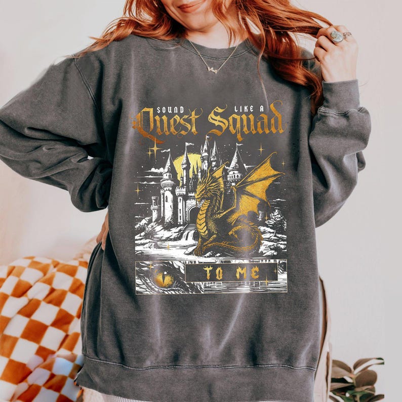 Op de afbeelding: Donkergrijze sweatshirt met een fantasie-afbeelding. Het ontwerp toont een kasteel, een gouden draak en de tekst "Sound like a Quest Squad to me" in gouden letters. De sweatshirt heeft lange mouwen.
