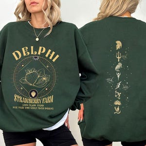May include: A dark green sweatshirt with a graphic of strawberries and the text "DELPHI STRAWBERRY FARM LONG ISLAND SOUND PICK YOUR OWN GODLY FRESH BERRIES". The back of the sweatshirt has a vertical row of gold symbols.