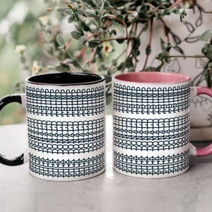 May include: Two ceramic mugs with a white base and a repeating black geometric pattern. One mug has a black interior and handle, the other has a pink interior and handle. The mugs are on a light surface.