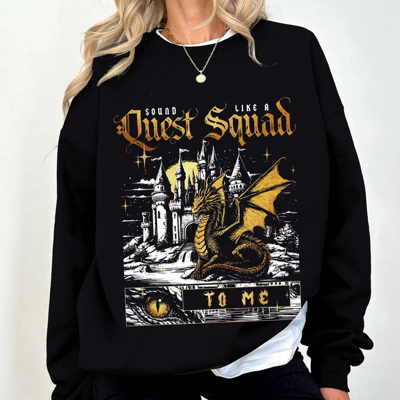 Op de afbeelding: Zwart sweatshirt met een fantasie-ontwerp. Het heeft een draak, kasteel en de tekst "Sound Like a Quest Squad To Me" in goud. De draak heeft gouden vleugels en staat voor een kasteel. Het ontwerp staat in een rechthoekige lijst.