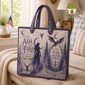 May include: A rectangular quilted tote bag in shades of purple and cream featuring a fantasy illustration of a person with a sword, a dragon, a deer, and text that reads: She was the heir of ash and fire and she would bow to no one.
