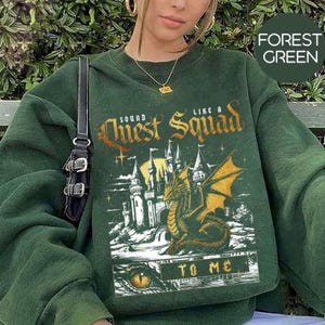 Op de afbeelding: Bosgroene sweatshirt met een fantasie-afbeelding. Het ontwerp toont een kasteel, een draak en de tekst "Sound Like a Quest Squad To Me". De sweatshirt heeft een effen bosgroene kleur.