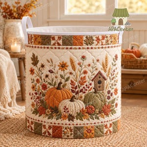 May include: A cylindrical quilted fabric storage basket featuring a cream-colored background with an embroidered autumn scene of orange, white, and green pumpkins, wheat stalks, flowers, and a birdhouse. The top and bottom borders have a patchwork design.