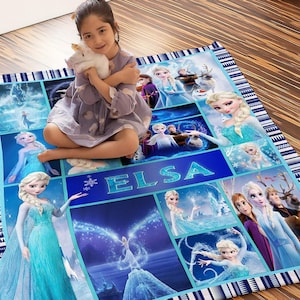May include: A blue and white patterned blanket featuring images of Elsa from the movie Frozen. The blanket has a blue border with white geometric designs and the name "ELSA" in large blue letters. A young girl is sitting on the blanket.