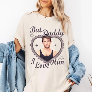 May include: Cream-colored t-shirt featuring a heart-shaped graphic with a man's portrait and the text "But Daddy I Love Him". The graphic is in shades of gray and black.