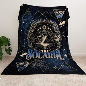 May include: A black fleece blanket with a gold and blue design. The design features a circle with the text "Zodiac Academy Solaria" and a starburst pattern. The blanket is perfect for cuddling up on the couch or in bed.
