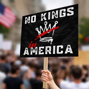 May include: A black sign with the words "NO KINGS" in white, above a white crown crossed out with a red line. Below the crown, the words "in AMERICA" are in red and white. The sign is held up in a crowd.