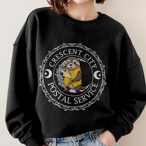 May include: Black sweatshirt with a white graphic of an otter holding a letter. The graphic is inside a circle with the text "Crescent City Postal Service" around it.