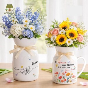 Puede incluir: Dos jarrones de cerámica blanca con arreglos florales. Uno tiene flores azules y blancas, el otro girasoles y rosas rosas. Ambos jarrones tienen una cinta color crema y el texto "Emma" y "Nana".