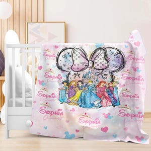 a pink and white blanket with a Disney princess theme, including a castle and various Disney characters. The blanket is placed on a wooden floor, and there is a white crib visible in the background.