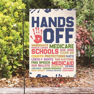 Könnte beinhalten: Eine Gartenflagge mit den Worten "Hands Off" in großen blauen Buchstaben. Die Flagge ist mit einem roten Handabdruck und einer Liste von Sozialprogrammen und Rechten verziert, darunter "Medicare", "Schools", "Clean Air", "LGBTQ+ Rights", "Medicaid" und "Social Security".