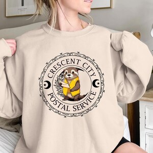 May include: A beige sweatshirt with a black and white graphic of an otter holding a letter. The graphic is a circular stamp with the text "Crescent City Postal Service".