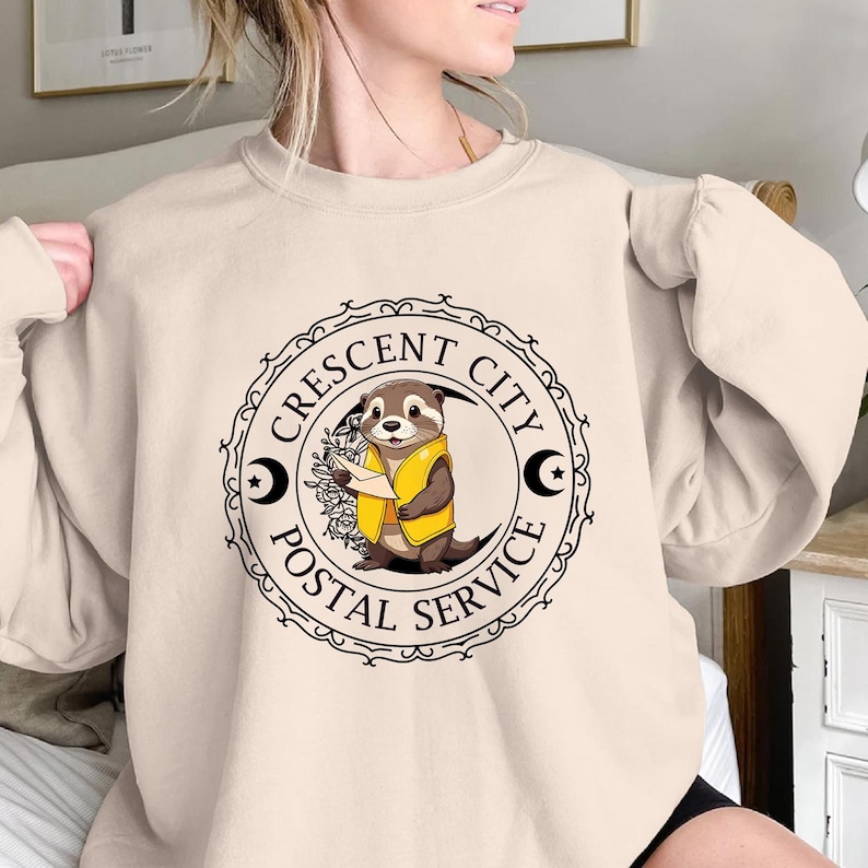 May include: A beige sweatshirt with a black and white graphic of an otter holding a letter. The graphic is a circular stamp with the text "Crescent City Postal Service".
