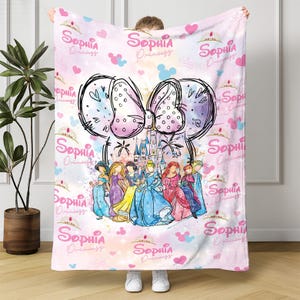 A person is holding a pink blanket with a Disney princess theme, featuring a group of princesses and a castle.