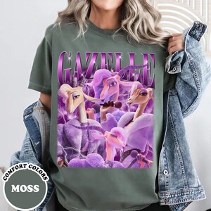 A person wearing a green t-shirt with a graphic design featuring purple and pink characters, including a unicorn and a bird, along with the text "Gazelle" in a stylized font.