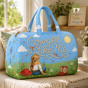 Puede incluir: Un bolso de viaje acolchado de color azul claro con una escena impresa de una persona con el cabello trenzado sentada en un campo. El texto "Cowgirl Like Me" est&aacute; escrito en una fuente estilo cuerda, junto a notas musicales, un sol y botas.