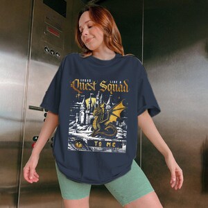Op de afbeelding: Marineblauw T-shirt met een afbeelding van een draak, kasteel en de tekst "Quest Squad Like a To Me". Het ontwerp is in tinten goud, wit en zwart. Het shirt wordt gedragen door een persoon die een groene short draagt.