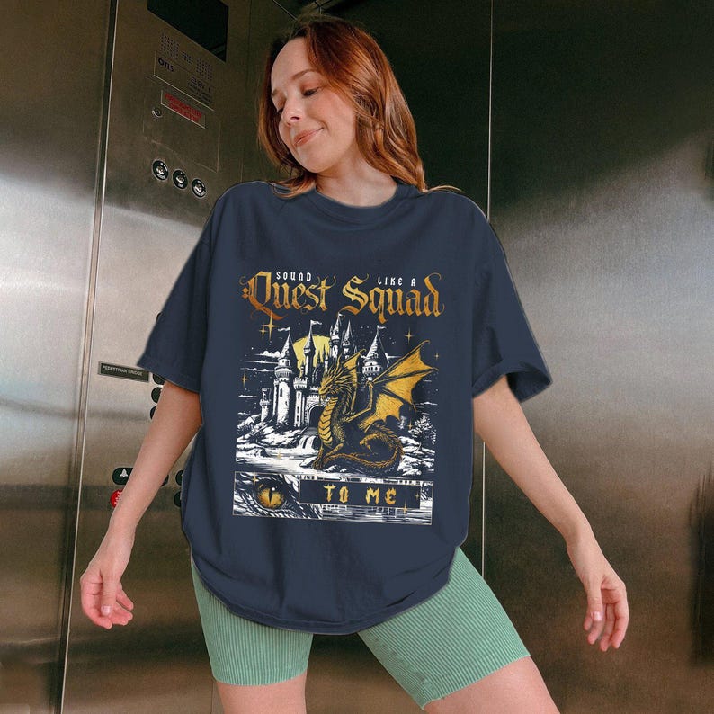 Op de afbeelding: Marineblauw T-shirt met een afbeelding van een draak, kasteel en de tekst "Quest Squad Like a To Me". Het ontwerp is in tinten goud, wit en zwart. Het shirt wordt gedragen door een persoon die een groene short draagt.