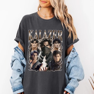 May include: Dark gray t-shirt featuring the word "KALAZER" in a metallic font above a collage of male portraits. The portraits are surrounded by lightning bolt graphics. The shirt is a casual, unisex style.