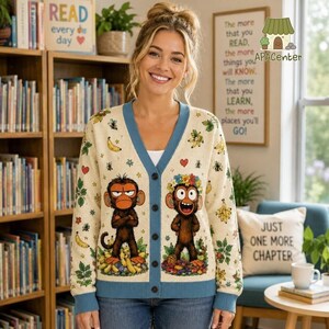 Könnte beinhalten: Eine cremefarbene Strickjacke mit V-Ausschnitt, blauem Saum und einem wiederkehrenden Muster aus Affen, Bananen und Blumen. Auf der Vorderseite befinden sich zwei große, unterschiedliche Affenillustrationen: einer mit mürrischem Gesichtsausdruck und einer mit einem Lächeln.
