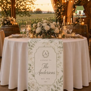 May include: A round table set for a wedding reception, featuring a white tablecloth and a floral table runner. The runner has a floral design with the text "Cheers to The Andersons EST. 2026". The table is decorated with flowers, candles, and glassware.
