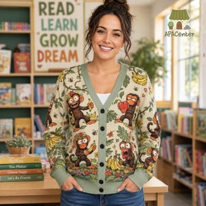 Könnte beinhalten: Eine cremefarbene Strickjacke mit V-Ausschnitt und einem durchgehenden Muster aus braunen Affen, gelben Bananen und grünem Blattwerk. Der geknöpfte Cardigan hat einen salbeigrünen Saum an Ausschnitt, Ärmelbündchen und Hüfte sowie fünf dunkle Knöpfe.