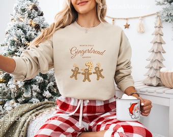 May include: A woman wearing a beige sweatshirt with the text "North Pole Gingerbread Bakery" and a graphic of three gingerbread men. She is also wearing red and white plaid pajama pants and holding a white mug with a Santa Claus design.