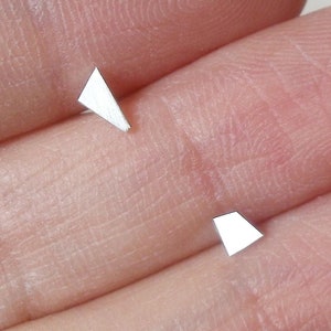 Op de afbeelding: Twee kleine, zilverkleurige, geometrische oorbellen. Eén oorbel heeft een driehoekige vorm, de andere een trapeziumvorm. De oorbellen worden op de vingers van een persoon getoond.