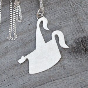 May include: A silver-colored necklace with a pendant shaped like a stylized ghost. The necklace chain is a delicate curb link style. The pendant is a solid, flat design with a matte finish. The necklace is displayed on a wooden surface.