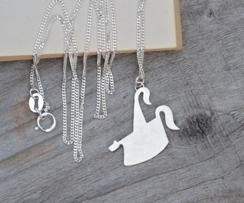 May include: A silver-colored necklace with a pendant shaped like a stylized ghost. The necklace chain is a delicate curb link style. The pendant is a solid, flat design with a matte finish. The necklace is displayed on a wooden surface.