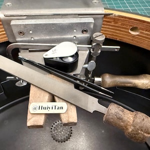 以下が含まれることがあります： 木製のハンドルが付いたジュエリー用ヤスリ、黒いジュエリー用ルーペ、黒いピンセットを備えた金属製のベンチピンとクランプのセット。@HuiyiTanというテキストが書かれた小さな長方形のタグが木製のベンチピンの上に置かれています。