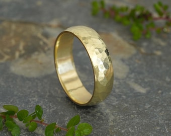May include: A gold ring with a hammered texture. The ring is a warm yellow color and has a rounded shape. The ring is sitting on a gray surface with green foliage in the background.
