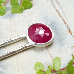 May include: A pair of silver hair sticks with a faceted ruby gemstone set in a silver bezel. The sticks have a hammered texture.